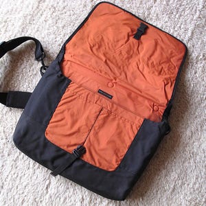 Op de afbeelding: Zwarte messenger bag met een oranje interieur. De tas heeft een zwarte, verstelbare schouderriem en een klepsluiting met een gesp. De binnenkant heeft een zak en een drukknoopsluiting.