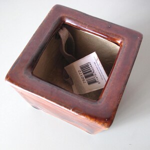 May include: A square, brown ceramic planter with a glossy finish. The interior is beige, and a tag with a barcode and numbers is inside. The planter is empty and sits on a white surface.