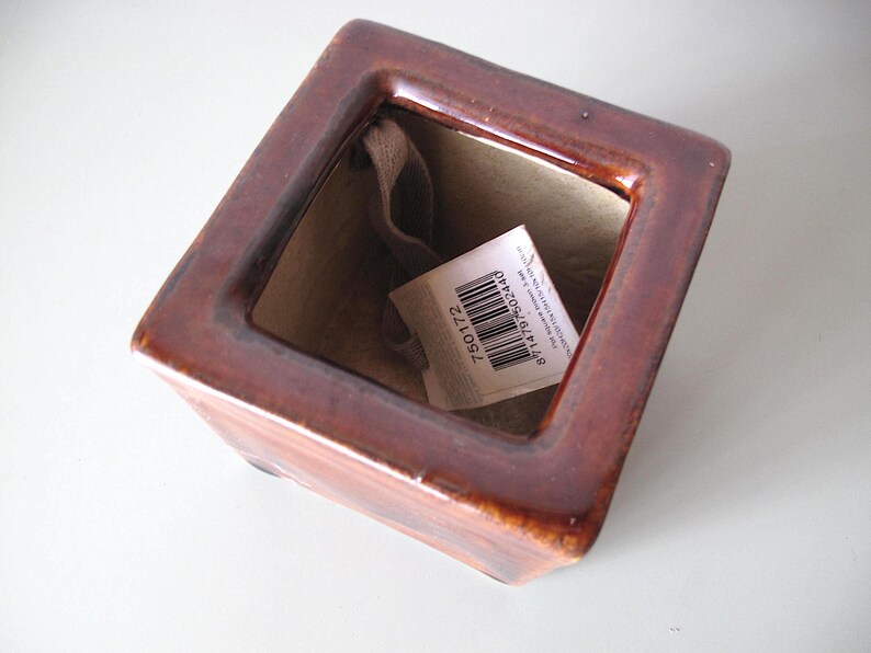 May include: A square, brown ceramic planter with a glossy finish. The interior is beige, and a tag with a barcode and numbers is inside. The planter is empty and sits on a white surface.