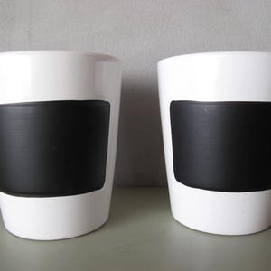 Könnte beinhalten: Zwei weiße Keramik-Blumentöpfe mit einem breiten schwarzen Band in der Mitte. Die Töpfe haben eine glatte, glänzende Oberfläche und ein schlichtes, modernes Design. Geeignet für den Innen- und Außenbereich.