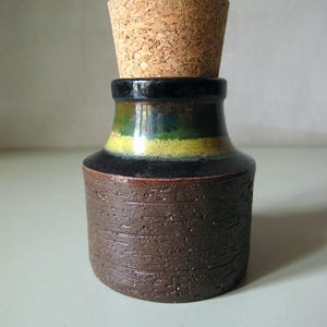 Pode incluir: Pequena garrafa decorativa de cerâmica com rolha de cortiça. A garrafa tem uma base castanha escura texturizada, uma secção intermédia preta e verde e uma rolha de cortiça. Provavelmente para armazenar líquidos ou como objeto decorativo.