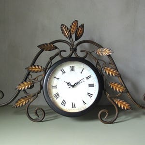 Può includere: Un orologio decorativo con cornice nera e quadrante bianco con numeri romani. L'orologio è adornato con foglie e volute in metallo color bronzo, creando un design elegante ispirato alla natura. L'orologio è progettato per essere posizionato su un tavolo o una mensola.