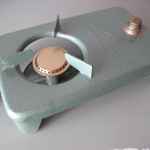 Peut inclure: Un réchaud de camping vintage, de couleur vert clair, avec un brûleur en laiton et un bouton de commande noir. Le réchaud a une base rectangulaire avec une ouverture circulaire pour le brûleur.