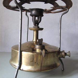 Pode incluir: Fogão de campismo de latão vintage com um topo circular e três pernas. O fogão tem um depósito de combustível de latão, um queimador e um botão de controlo. O topo tem um design circular com recortes. Projetado para cozinhar ao ar livre.