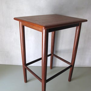 Könnte beinhalten: Ein hoher, quadratischer Holzhocker mit einer satten, rotbraunen Oberfläche. Der Hocker verfügt über vier schlanke, zylindrische Beine, die an allen vier Seiten durch horizontale Stützstangen verbunden sind und eine stabile Struktur bilden.