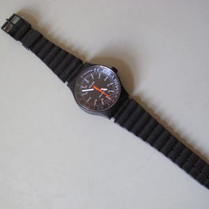 Könnte beinhalten: Schwarze Armbanduhr mit rundem Zifferblatt und schwarzem, geripptem Armband. Das Zifferblatt der Uhr hat weiße und orangefarbene Markierungen. Das Armband besteht aus flexiblem Material und wird mit einer schwarzen Schnalle befestigt.