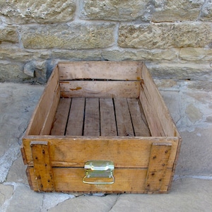 Könnte beinhalten: Eine verwitterte Holzkiste mit Metallverschluss und Griff. Die Kiste ist aus hellem Holz gefertigt und hat einen Lattenboden. Die rechteckige Kiste scheint aus der Vintage-Ära zu stammen.