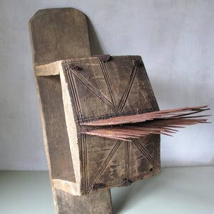 Op de afbeelding: Een verweerd houten object met een rechthoekige doos met gesneden geometrische patronen en metalen beslag, bevestigd aan een lange, platte basis. Dunne houten spijkers steken uit de zijkant. Het lijkt een antiek gereedschap of decoratief item te zijn.