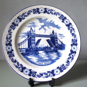 May include: A round ceramic plate featuring a blue and white transferware design. The center depicts Tower Bridge with the text TOWER BRIDGE below it. The rim is decorated with a repeating pattern of blue flowers, leaves, and acorns on a white background.