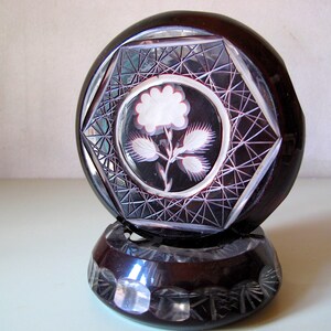 May include: A dark brown cut glass paperweight with a floral design. The top section features a white flower and leaves within a geometric pattern. The base is a smaller, matching piece with a similar cut glass design.