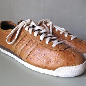 Puede incluir: Un par de zapatillas de cuero marrón con cordones y suelas blancas. Los zapatos presentan el diseño clásico de tres rayas en los laterales y una superficie texturizada. Las zapatillas son de estilo atlético clásico.