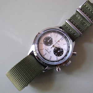 May include: A silver wristwatch with a cream-colored face, black sub-dials, and a green nylon strap. The watch face features the text "Zhi Yuan" and a red star. The strap is approximately 0.75 inches wide.