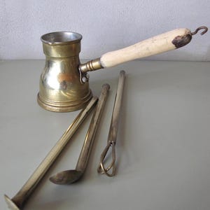 Könnte beinhalten: Eine Kaffeekanne aus Messing mit einem hellen Holzgriff und einem Haken. Drei silberfarbene Metallwerkzeuge sind vor der Kanne angeordnet. Die Kanne hat einen zylindrischen Körper und einen Ausguss.