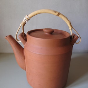 May include: A terracotta teapot with a curved spout and a lid with a small knob. The handle is made of woven light brown material. The teapot is a warm reddish-brown colour.