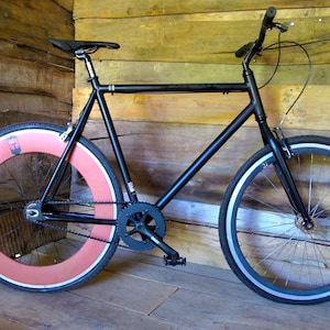 Könnte beinhalten: Ein schwarzes Fixed-Gear-Fahrrad mit rotem Hinterrad und schwarzem Vorderrad. Das Fahrrad hat einen schwarzen Rahmen, Sattel und Lenker. Die Reifen haben einen silbernen Streifen. Das Fahrrad lehnt an einer Holzwand.