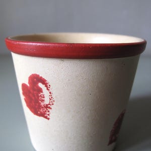 May include: A small, off-white ceramic pot with a red rim and red heart-shaped designs. The pot has a slightly textured surface. Suitable for small plants or as a decorative item. The pot's diameter is approximately 7 cm.