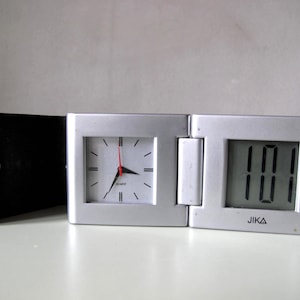 Op de afbeelding: Een zilveren opvouwbare reisklok met een vierkante analoge wijzerplaat en een digitale display. De analoge klok heeft zwarte wijzers en markeringen met een rode secondewijzer. De digitale display toont de tijd. De merknaam "JIKA" is afgedrukt.