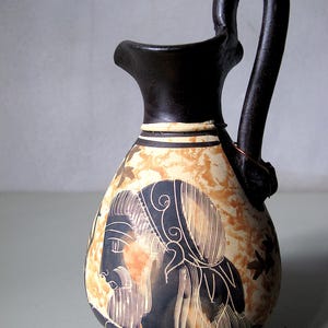 Może przedstawiać: Ceramiczny dzban z czarnym uchwytem i wylewką. Dzban ma kremowy korpus z brązowym i beżowym wzorem oraz czarną ilustrację profilu osoby.