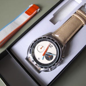 May include: A wristwatch with a cream-colored canvas band and a black and white face. The watch face features a small dial and orange and blue hands. An additional watch band with white, blue, and orange stripes is also included.