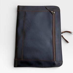 May include: A dark blue leather portfolio with a brown zip and stitching. The portfolio has a rectangular shape with rounded corners and a leather pull tab on the zip. The stitching is visible along the edges and on the front.