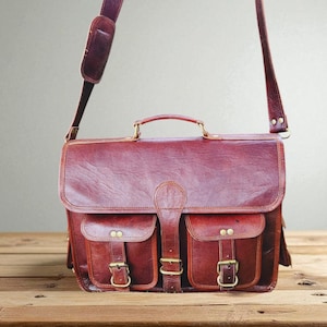 May include: A burgundy leather messenger bag with two front pockets and brass-colored buckles. The bag has a top handle and a long, adjustable shoulder strap. The bag is sitting on a wooden surface.