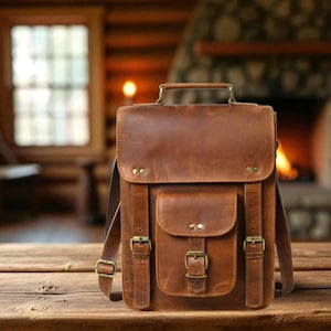 Pode incluir: Uma mochila de couro castanho com uma pega superior, bolso frontal e alças ajustáveis. A bolsa tem fivelas e rebites de cor latão. O fundo apresenta uma mesa de madeira e uma lareira.
