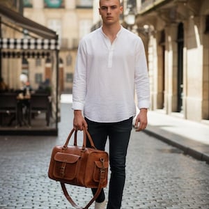 Puede incluir: Una persona con una camisa blanca de manga larga y vaqueros negros, que lleva una bolsa de viaje de cuero marrón con una correa para el hombro. La bolsa tiene dos bolsillos delanteros y herrajes metálicos. La persona camina por una calle adoquinada.