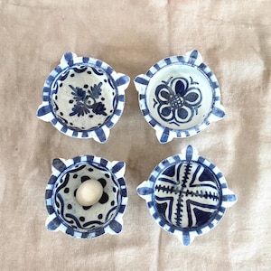 May include: Four small, round ceramic bowls with a white and blue floral design. Each bowl has a unique pattern, with a mix of flowers, dots, and geometric shapes. The bowls have a scalloped edge and are arranged on a neutral surface.