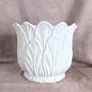 May include: A white ceramic planter featuring a sculpted exterior with overlapping leaf patterns that create a scalloped rim. The vessel has a rounded body and a circular base, with a glossy finish that highlights the intricate botanical texture.