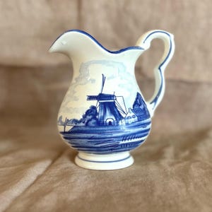 May include: A small, white ceramic pitcher with a blue windmill and landscape design. The pitcher has a curved handle and a spout. The design features a windmill, trees, and water, all in shades of blue.