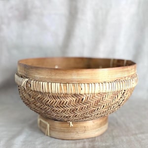 May include: A round, footed bowl crafted from woven light brown bamboo strips. The bowl features a complex herringbone weave pattern on the sides, a smooth upper rim, and a solid, circular base, all held together with thin, light-colored binding.