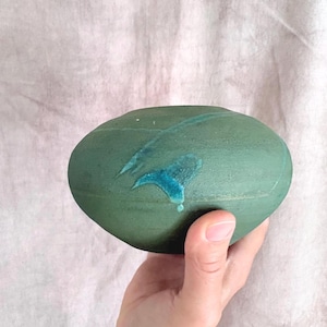 May include: A rounded, matte-finish ceramic bowl with a deep forest green base color. The surface features a distinct, glossy turquoise blue glaze accent that streaks across the center, creating a contrast in texture and color on the object.