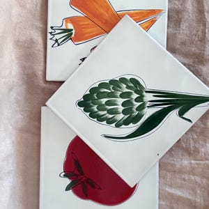 May include: Three square white ceramic tiles featuring hand-painted vegetable illustrations. One tile displays two orange carrots, another shows a green artichoke, and the third depicts a single red tomato, all outlined in dark blue ink.