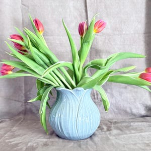 May include: A light blue ceramic vase shaped like a tulip, filled with vibrant pink and yellow tulips with green leaves. The vase has a textured, petal-like design. The flowers are arranged to create a full, eye-catching display.