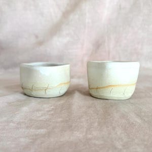 May include: Two small, off-white ceramic cups with a matte finish. Each cup features a subtle, light brown horizontal band near the base, accented by a series of delicate, evenly spaced vertical etched lines encircling the lower exterior.