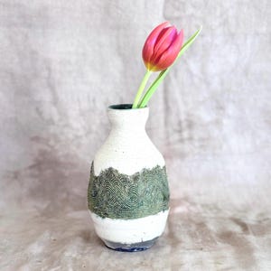 May include: A white ceramic vase with a textured green wave pattern around the middle. A pink and yellow tulip with green stem and leaf is placed inside the vase. The vase has a dark green interior and a blue base.