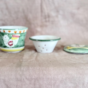 May include: A ceramic teacup, small bowl, and lid set. The teacup and lid are green with a bird design. The bowl is white with small brown dots. The set is on a pink surface.