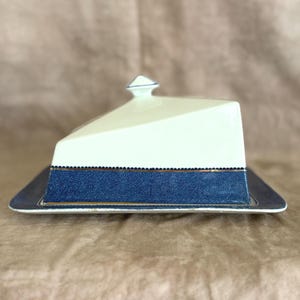 May include: A rectangular butter dish with a white, triangular lid and a navy blue base. The base has a gold trim and a textured appearance. The lid has a small, triangular finial with blue accents.