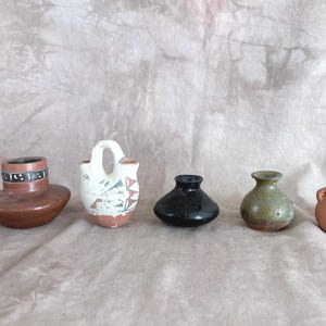 May include: A collection of five small ceramic vessels including a wide brown pot with a patterned rim, a white double-spout vessel with geometric designs, a black rounded pot, a mottled green vase, and a small plain terracotta-colored jar.
