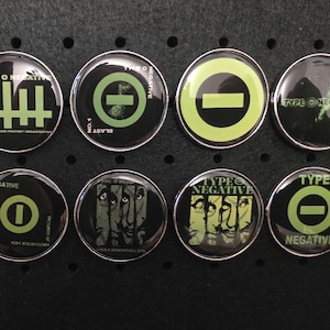 Könnte beinhalten: Eine Sammlung von acht runden Buttons der Band Type O Negative. Die Buttons zeigen verschiedene Designs, darunter das Logo der Band, Gesichter und Text. Das Farbschema ist hauptsächlich Schwarz und Grün, mit silbernen Rändern.