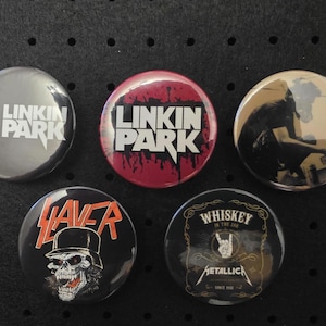 Könnte beinhalten: Eine Sammlung von fünf runden Metall-Ansteckbuttons mit Bandlogos und -bildern. Die Buttons zeigen Designs für Linkin Park, Slayer und Metallica. Die Farben sind unter anderem Schwarz, Rot und Silber. Ein beliebtes Accessoire für Musikfans.