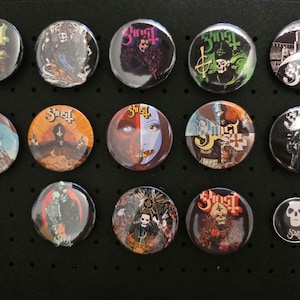 Könnte beinhalten: Eine Sammlung von fünfzehn runden Buttons mit verschiedenen Designs und dem Wort "Ghost" in gotischer Schrift. Die Buttons zeigen Bilder von Totenköpfen, religiösen Figuren und anderen gotischen Bildern. Die Buttons sind auf einer schwarzen Oberfläche angeordnet.