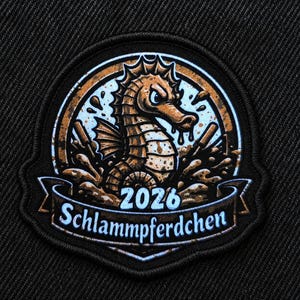 Könnte beinhalten: Ein kreisförmiger bestickter Aufnäher mit einem braunen Seepferdchen, das aus Schlammspritzern auftaucht. Das Design enthält den Text 2026 und Schlammpferdchen in Hellblau auf einem Banner unten, alles auf schwarzem Grund mit gesticktem Rand.