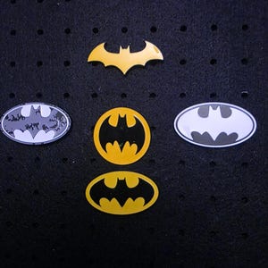 Könnte beinhalten: Eine Sammlung von Batman-Logo-Anstecknadeln in verschiedenen Formen und Farben. Die Anstecknadeln umfassen ein gelbes Fledermaussymbol sowie ovale und kreisförmige Designs mit gelben und schwarzen oder schwarz-weißen Farbschemata. Die Anstecknadeln werden auf einer schwarzen Oberfläche präsentiert.