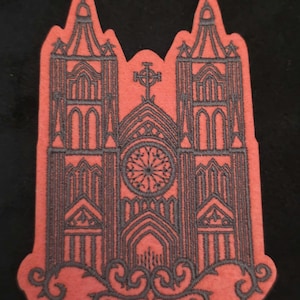 Könnte beinhalten: Ein korallenfarbener Stoffaufnäher mit dunkelgrauer Stickerei einer Kathedrale mit zwei hohen Türmen, einem zentralen Rosenfenster und einem Kreuz an der Spitze. Die Basis des Aufnähers ist mit komplizierten, geschwungenen Ornamenten verziert.