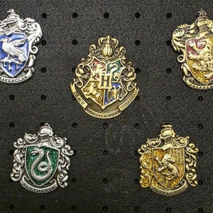 Könnte beinhalten: Fünf Metallwappen mit den Emblemen der Hogwarts-Häuser: Ravenclaw, Gryffindor, Slytherin, Hufflepuff und das Hogwarts-Wappen. Jedes Wappen hat aufwendige Details und leuchtende Farben, vor einem schwarzen Hintergrund.