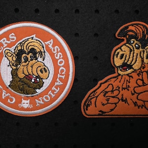 Könnte beinhalten: Zwei aufgestickte Aufnäher mit der Figur Alf. Einer ist ein runder Aufnäher mit dem Text "CAT LOVERS ASSOCIATION" und einem Bild von Alf. Der andere Aufnäher ist ein Bild von Alf mit erhobenen Händen.