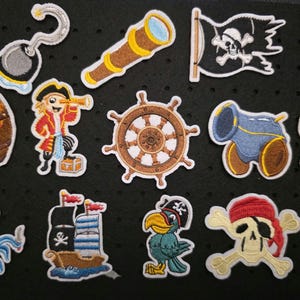 Könnte beinhalten: Eine Sammlung farbenfroher, gestickter Piratenaufnäher. Enthält ein Piratenschiff, Totenkopf und gekreuzte Knochen, eine Schatzkiste, einen Papagei, eine Kanone, ein Fernrohr, ein Steuerrad und einen Piraten mit einem Teleskop.