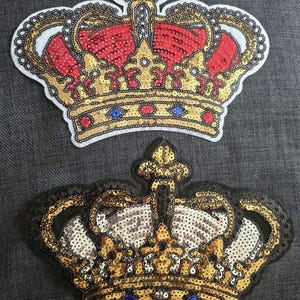 Könnte beinhalten: Zwei dekorative Kronenaufnäher. Die obere Krone hat rote, goldene und blaue Pailletten mit weißem Hintergrund und goldenem Rand. Die untere Krone hat goldene und schwarze Pailletten mit weißem Hintergrund und goldenem Rand. Beide Kronen haben ein königliches Design.