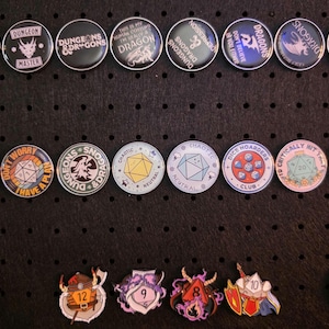Könnte beinhalten: Eine Sammlung runder Anstecknadeln und Buttons mit verschiedenen Designs und Texten, darunter Drachen, Würfel und Sätze wie "Dungeon Master". Die Pins sind auf einer schwarzen Oberfläche angeordnet.